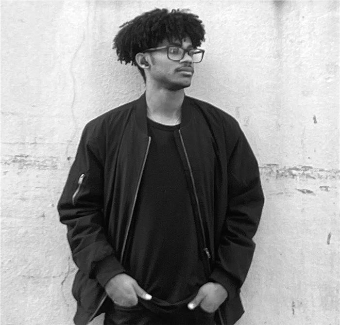 Jules standing against a weathered white wall, wearing a black bomber jacket